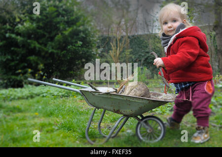 Baby Mädchen im roten Mantel in einem Garten mit einer Schubkarre arbeiten Stockfoto