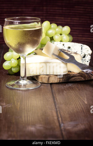 Milchglas mit Weißwein, frischen Trauben und verschiedene Arten von Käse mit einer Gabel auf einem Holztisch stehen. Stockfoto