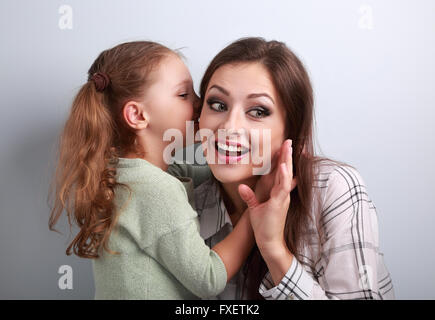 Glückliches Kind Mädchen flüsterte das Geheimnis zur Mutter überraschend ins Ohr mit lustigen Gesicht auf blauem Hintergrund Stockfoto