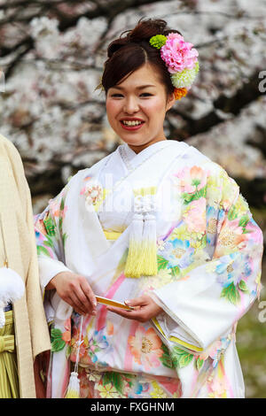 Lächelnden jungen Frau, Japanisch braut, im Kimono und Holding geschlossen Ventilator, stehend, mit unscharfen Kirschblüte im Hintergrund posiert. Frühling. Stockfoto