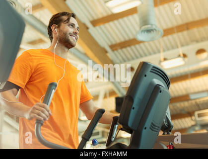 Lächelnder Mann mit Kopfhörern mit Crosstrainer im Fitnessstudio Stockfoto