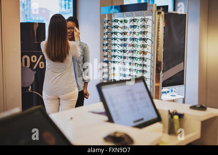 Saleslady Unterstützung eines Kunden in einem Geschäft versuchen auf Brillen mit Blick auf einen Computer im Vordergrund. Stockfoto