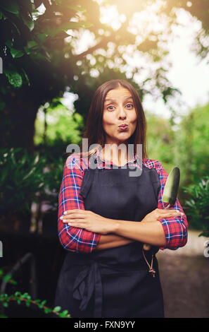 Frech verspielte junge Frau schmollend ihre Lippen in die Kamera, während sie im Freien im Garten mit einer Kelle in ihre Hände unter steht eine Stockfoto