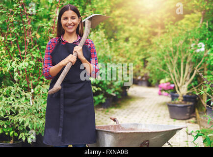 Glückliche junge Frau arbeitet in einer kommerziellen Gärtnerei halten einen Spaten über die Schulter, während sie unter den Pflanzen mit steht ein whee Stockfoto