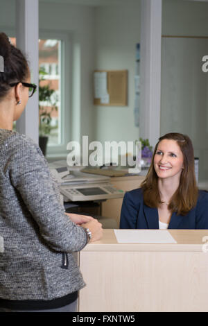 Lächelnde Angestellte (Kaukasier, im Hintergrund) sitzt hinter einer Theke und sucht für einen Kunden (lateinisch, im Vordergrund) Stockfoto