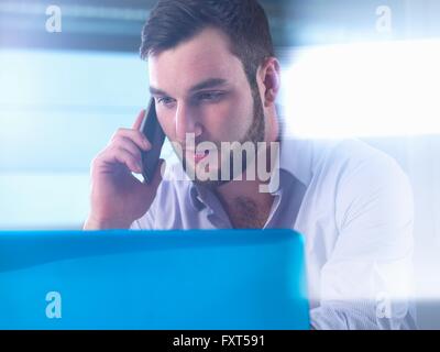Junger Geschäftsmann auf seinem Smartphone sprechen während der Arbeit an Laptop im Büro Stockfoto