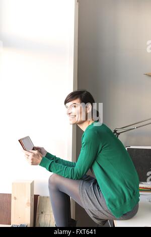 Seitenansicht des Reife Frau sitzen am Schreibtisch Holding Digital tablet aussehende Weg Lächeln Stockfoto