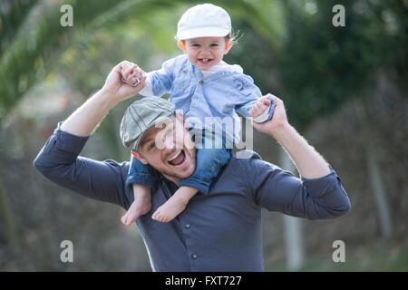 Baby Boy mit Baseballmütze sitzt auf Vaters Schultern Blick auf die Kamera zu Lächeln Stockfoto