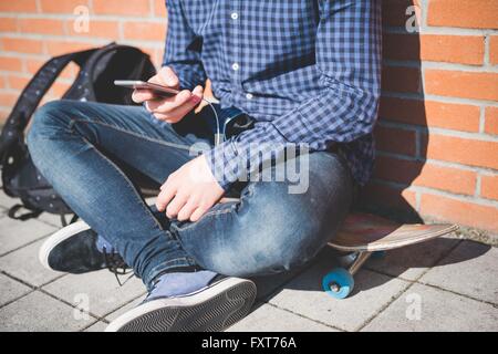 Hals abwärts Blick auf junge männliche urban Skateboarder sitzen auf Bürgersteig Smartphone Text zu lesen Stockfoto