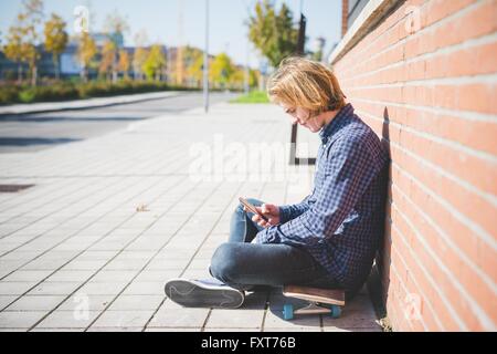 Junge männliche urban Skateboarder sitzen auf Bürgersteig Auswahl Smartphone Musik Stockfoto