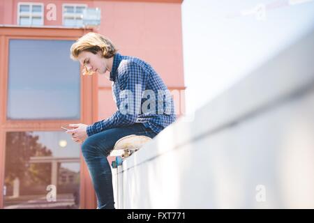Junge männliche urban Skateboarder sitzen auf Skateboard Smartphone Text zu lesen Stockfoto