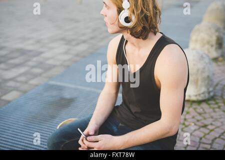 Junger Mann sitzt am Bürgersteig Kopfhörer Musik hören Stockfoto