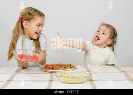 Zwei kleine Mädchen an einem Tisch vorbereitet Pizza. Eine mit skurrilen Blick erstreckt sich eine Hand auf einen Teller mit Tomaten, führt die Stockfoto