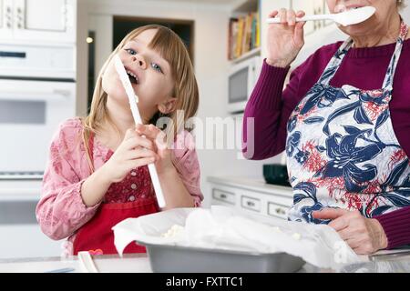 Mädchen und Oma lecken Kuchen-mix vom Spatel am Küchentisch Stockfoto