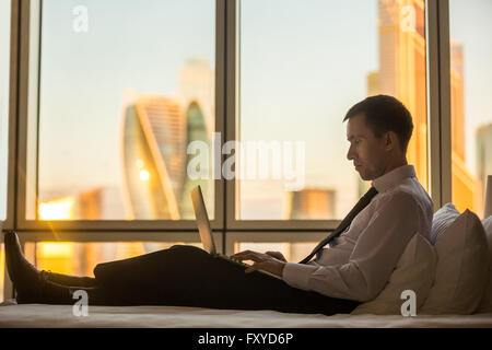 Geschäftsmann, arbeiten von zu Hause oder auf Reise. Seite Ansicht Porträt von hübscher junger Geschäftsmann auf Bett sitzt und mit laptop Stockfoto
