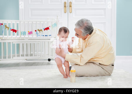 Ältere Mann kümmert sich um ein Baby mit hält das Baby sitzt auf dem Boden mit einer Babyflasche Stockfoto