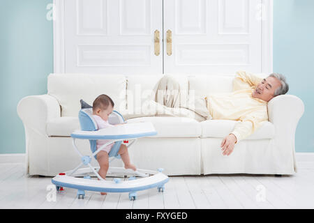 Müde senior Mann auf einer Couch liegen und Blick auf ein Baby auf eine Lauflernhilfe Stockfoto