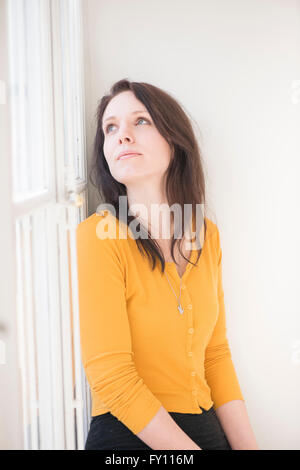 Frau sitzt durch Fenster, Blick nach oben. Lifestyle-Bild zeigt eine selbstbewusste Frau denken. Konzept der Kontemplation. Stockfoto