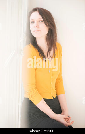 Frau sitzt durch Fenster, Blick in die Kamera. Lifestyle-Bild zeigt eine selbstbewusste Frau denken. Konzept der Kontemplation. Stockfoto