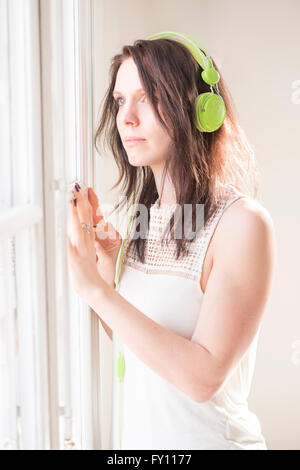 Frau am Fenster stehend, wegschauen. Sie ist im grünen Kopfhörer Musik hören. Lifestyle-Bild der Kontemplation. Stockfoto
