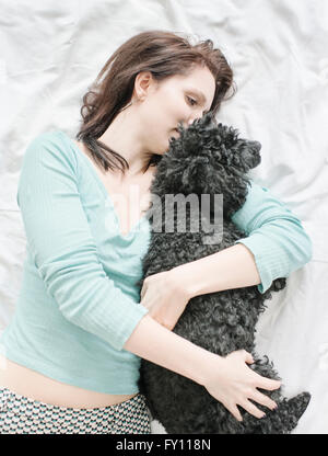 Frau liegend im Bett umarmt schwarzen Pudel. Lifestyle-Bild zeigt Zuneigung und die Bindung zwischen Hund und Mensch. Stockfoto