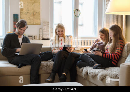 Glückliche Eltern mit Töchtern mit Laptop und mobilen Geräten im Wohnzimmer Stockfoto