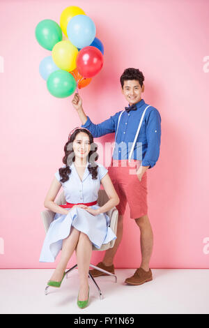 Junge lächelnde Frau im retro-Stil Kleid sitzen auf Stuhl und lächelnde Jüngling im Retro-Stil stehen mit Luftballons Stockfoto
