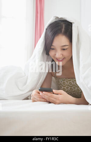 Porträt des jungen lächelnde Frau mit Smartphone unter einer Decke im Bett Stockfoto
