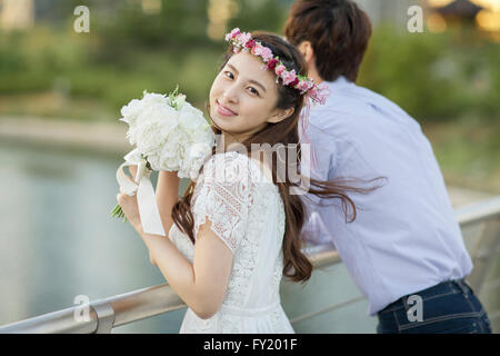 Braut in Blume Krone und weißes Kleid auf einer Brücke hält einen Blumenstrauß neben den Bräutigam Stockfoto