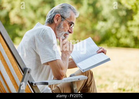 Alte Mann liest ein Buch auf dem Liegestuhl im Park im Sommer Stockfoto