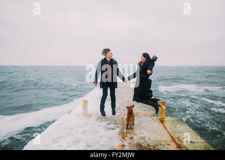 Romantischer Rückansicht eines jungen Paares zusammen mit kleinen Hund wandern und Spaß auf stoned Pier während regnerischen Herbsttag. Winter-Meer-Hintergrund Stockfoto