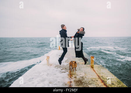 Romantischer Rückansicht eines jungen Paares zusammen mit kleinen Hund wandern und Spaß auf stoned Pier während regnerischen Herbsttag. Winter-Meer-Hintergrund Stockfoto