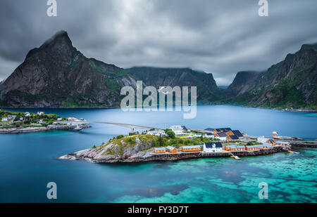 Mount Olstind über dem gelben Kabinen und dem türkisfarbenen Wasser des Sakrisoy Fischerdorf auf Lofoten in Norwegen am Abend bl Stockfoto