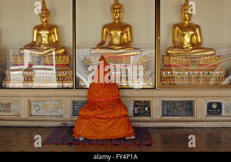 Eine neue buddhistische Statue ist in Orange gehüllt, die Roben warten vor drei glänzenden gold Buddahs sitzt Stockfoto