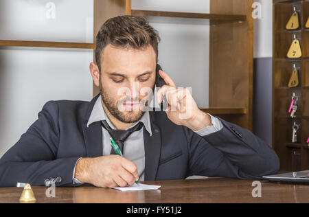 Porträt der Empfangsdame Notiz auf ein Stück Papier zu nehmen, während er an seinem Schreibtisch in einem kleinen Hostel am Telefon ist. Stockfoto