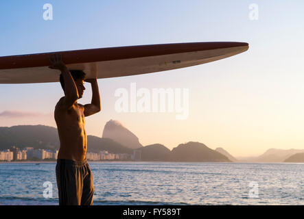 RIO DE JANEIRO - 5. April 2016: Ein junger brasilianischen Mann am Strand der Copacabana gleicht einem Surfbrett auf dem Kopf bei Sonnenaufgang. Stockfoto