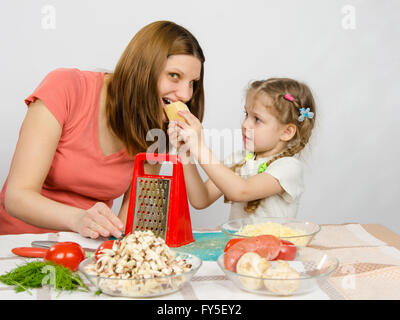 Sechsjährigen Mädchen mit Zöpfen geben Mama einen Bissen von einem Stück Käse am Küchentisch wo sie gemeinsam kochen Stockfoto