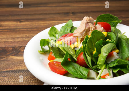 Frischer Spinat-Salat mit Thunfisch und Mais, kleine Stücke von Kirschtomaten in weißem Teller auf Holztisch Stockfoto