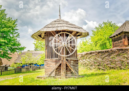 Alte hölzerne Wasser gut Haus mit einem großen Rad in der Nähe von einem gewebten Weide Zaun mit grünen Bäumen im Hintergrund. Stockfoto