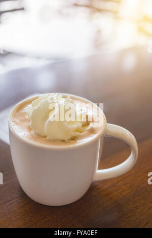 Fokus auf heißen Mokkakaffee mit Milch-Sahne in weißen Tasse auf Holztisch und helles Licht in der Nähe Stockfoto