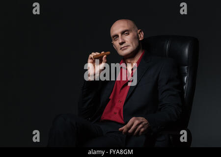Sicher gut aussehend Geschäftsmann sitzen im Bürostuhl und rauchende Zigarre auf schwarzem Hintergrund Stockfoto