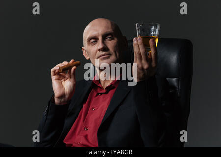 Attraktiver Geschäftsmann, raucht Zigarre und Whisky im Bürostuhl über schwarzem Hintergrund genießen Stockfoto
