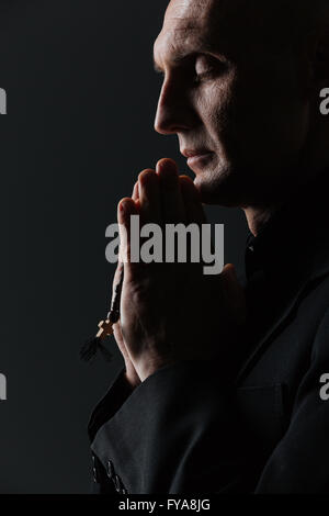 Mann mit geschlossenen Augen halten Rosenkranz und beten auf schwarzem Hintergrund Stockfoto