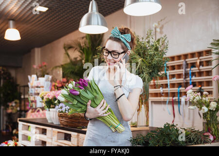 Fröhliche attraktive junge Frau Floristen halten zwei Trauben von Tulpen und telefonieren mit Handy im Blumenladen Stockfoto