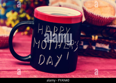 der Satz glücklich Muttertag handschriftlich mit Kreide in eine schwarze Tasse mit Kaffee, mit einigen Muffins im Hintergrund in einem Set t Stockfoto