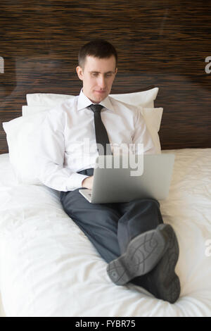 In voller Länge Portrait von Ernst hübscher junger Mann in Abendgarderobe auf Kissen auf dem Bett zu sitzen und zu Hause arbeiten Stockfoto