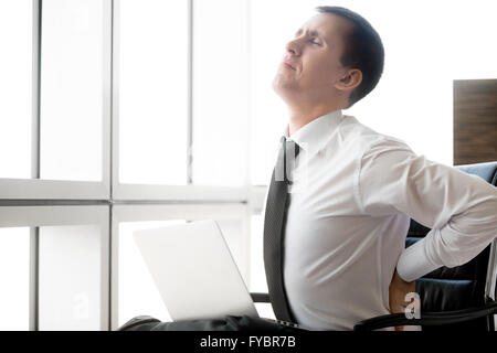 Hübscher Junge betonte Geschäftsmann sitzt in seinem Büro mit Laptop. Kaukasische Unternehmer leiden unter Rückenschmerzen Stockfoto
