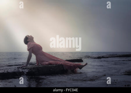eine Frau in einem rosa Kleid ist auf den Steinen am Meer liegen. Stockfoto