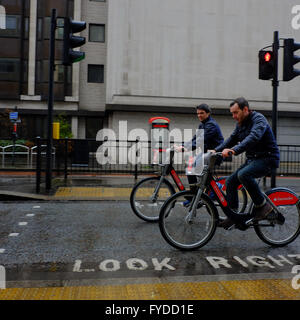 zwei Männer reiten Boris Bikes im Regen über "Look Right" Markierung unterwegs in London Stockfoto