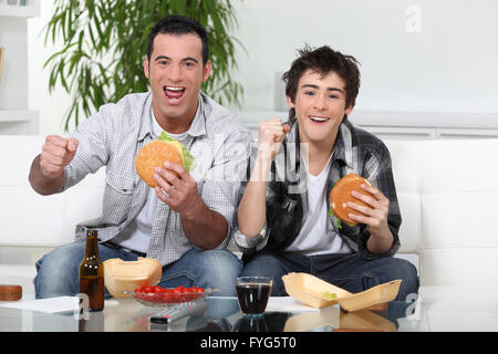 Vater und Sohn essen Burger vor Fernseher Stockfoto